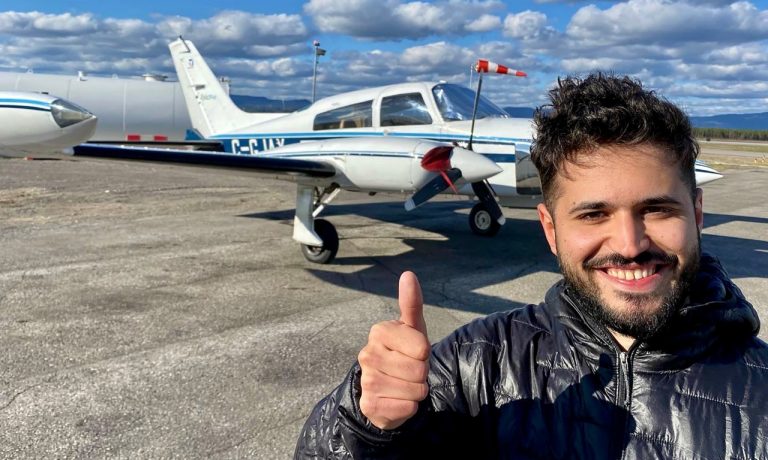 Young man in a puffer jacket, standing in front of a small propeller plane and giving a thumbs up.