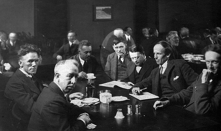 A black and white archival image of seven men in suits sitting at a restaurant table