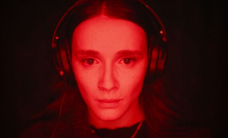 A young woman staring into a screen behind the camera, wearing headphones, and with a red light wash over her face.