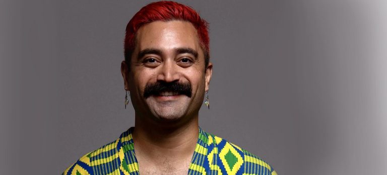 Smiling man with short, dyed red hair, a dark brown moustache and wearing a printed shirt.