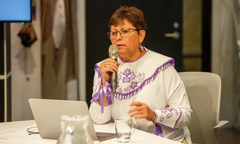 A woman with short, dark hair, wearing glasses and a white tunic with purple embroidery, sitting at a table and speaking into a microphone.