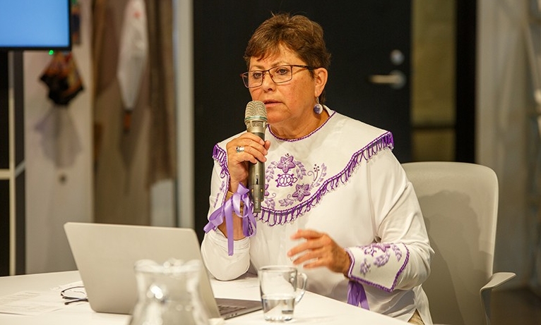 Donna Kahérakwas Goodleaf, director of decolonizing curriculum and pedagogy A woman with short, dark hair, wearing glasses and a white tunic with purple embroidery, sitting at a table and speaking into a microphone.