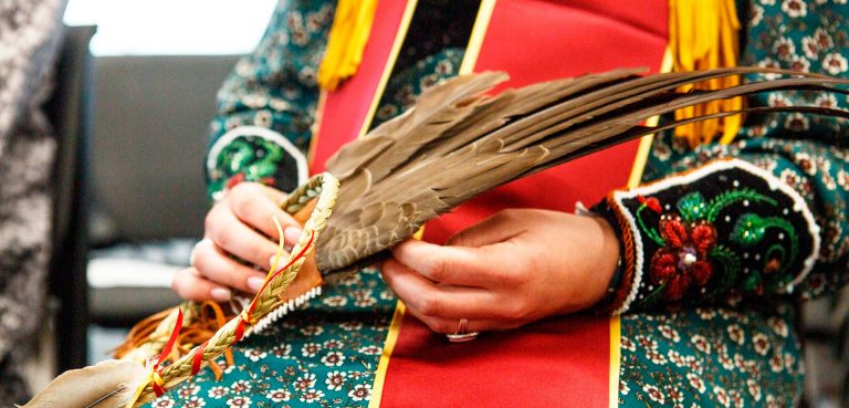 The hands of a person holding a large brown feather, wearing a patterned dress and a graduation sash.