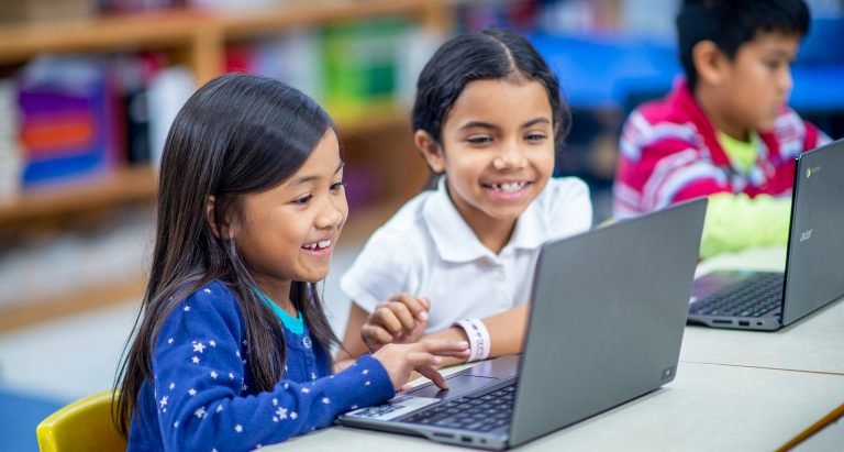 Young children sitting in a classroom setting and working on a laptop computer