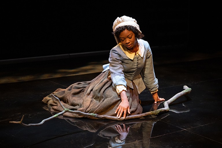 A young woman in period clothing — a bonnet and full length dress — kneeling on a stage.