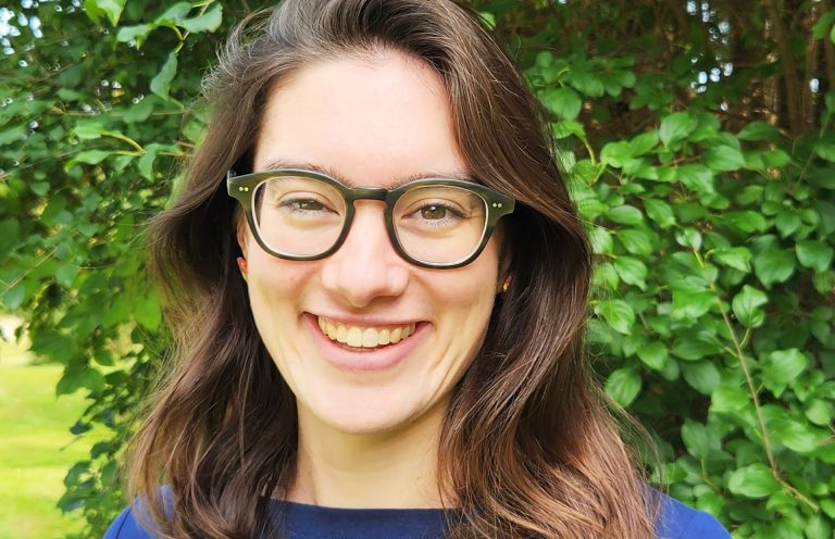 Smiling woman with long, light brown hair and glasses, wearing a blue shirt and outdoors