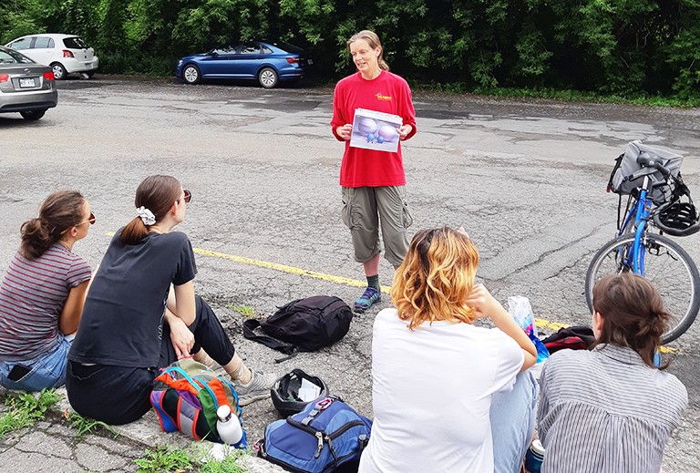 Karen Fisher Favret demonstrates a day camp activity to the FUN team and Concordia student volunteers just outside of the Falaise Saint-Jacques main entrance. | Photo: Ashley Spanier-Levasseur A group of young men and women sit on the curb and listen to an older woman doing a presentation