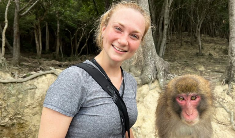 Young, blonde woman with a grey T-shirt standing in an outdoor setting and with a pink-faced Japanese macaque monkey beside her.