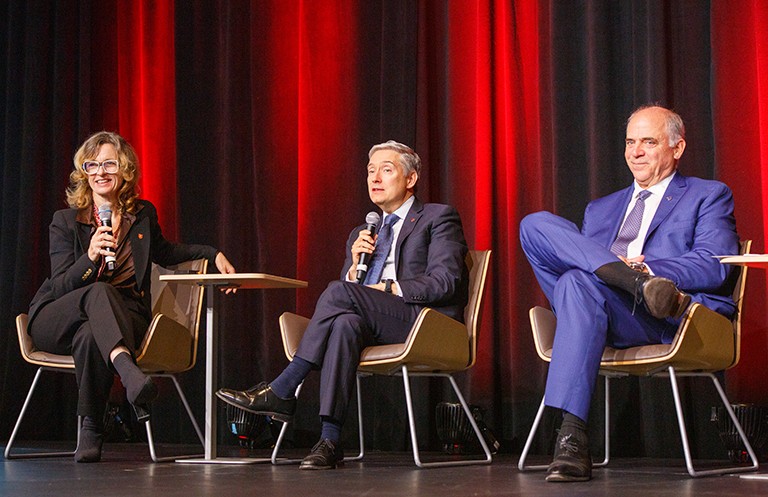 A panel of three — a woman and two men — seated on a stage and speaking into microphones