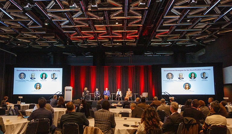 Photo of a large room filled with chairs and round tables, with a stage and panel of speakers at the front.