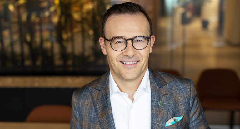 Smiling man with short, dark hair, wearing tortoise-shell-rimmed glasses, a suit shirt and a blazer.