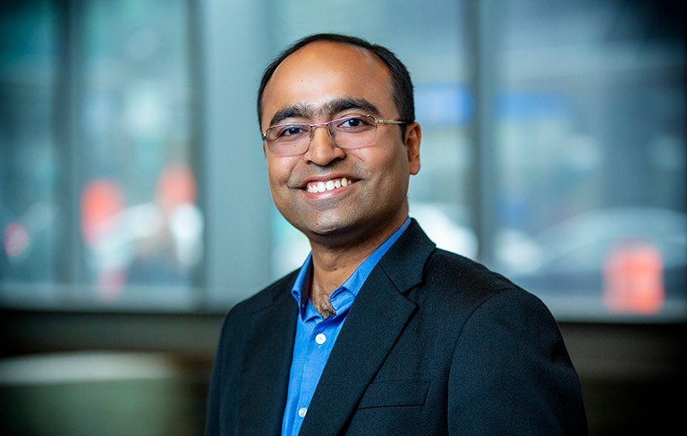 Smiling man with short, black hair, wearing glasses, a blue shirt and dark blue blazer.