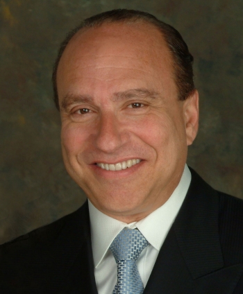 Smiling man with smoothed back short, dark hair, wearing a dark suit and blue tie