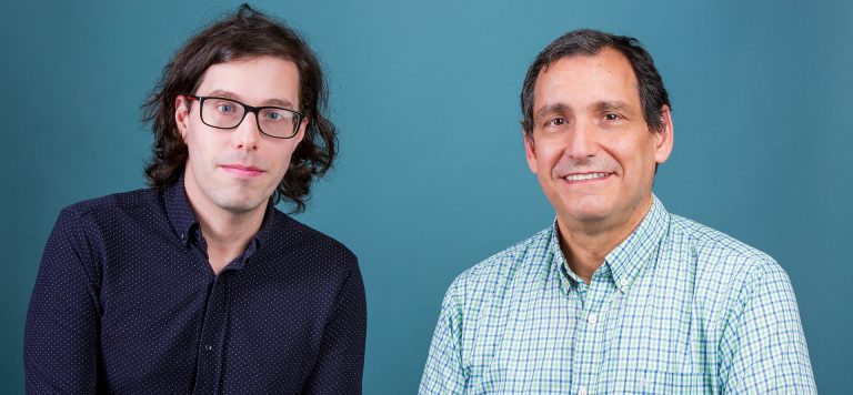 Two men, one younger with long hair and glasses, the other older with short, dark hair, and both wearing collared shirts.