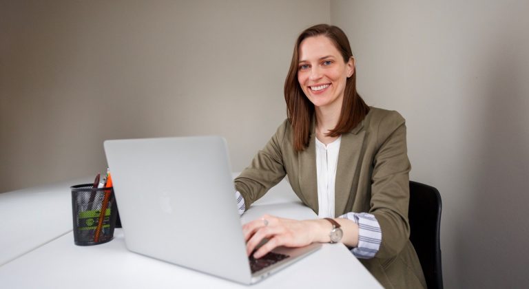 Rachael Lindberg sits at a computer wearing a white shirt and brown jacket