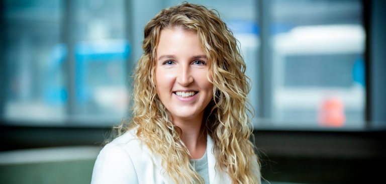 Smiling young woman with long, curly blonde hair, wearing a grey top and a white blazer.