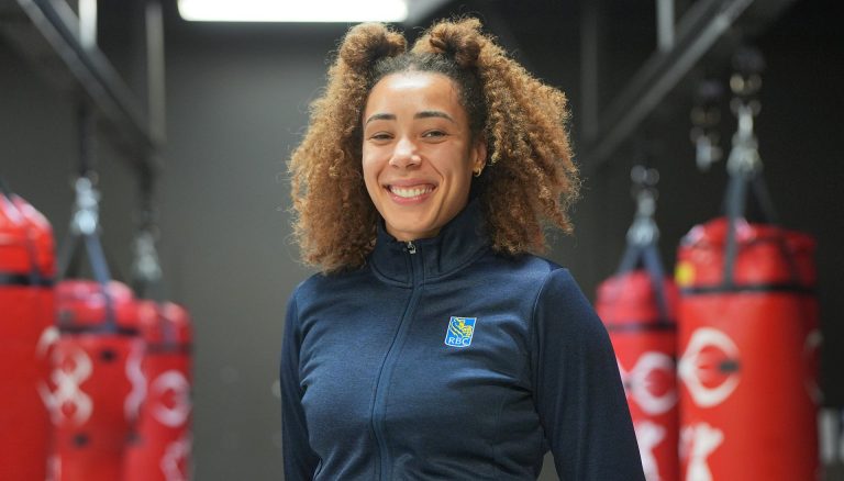 Smiling woman with long, curly hair, standing in a gym with many hanging boxing bags.
