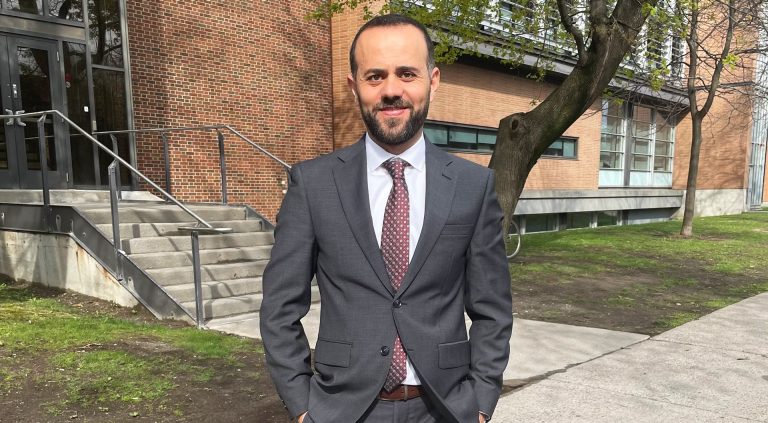 Smiling man with short, dark hair and a beard, wearing a grey suit and dark-red tie, and standing outside a university building.