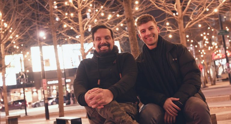 Two smiling men sitting on a bench in a downtown city area with Christmas lights in the trees in the background.