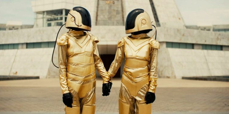 Two figures in golden space suits with helmets and dark visors, looking at one another and holding hands.