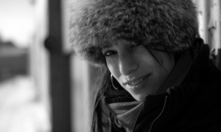 Black and white photo of a smiling woman wearing a scarf and a large feathered winter hat
