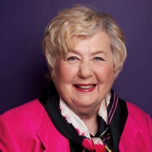 Smiling woman short(ish) blonde hair, wearing red lipstick and a bright pink blazer.