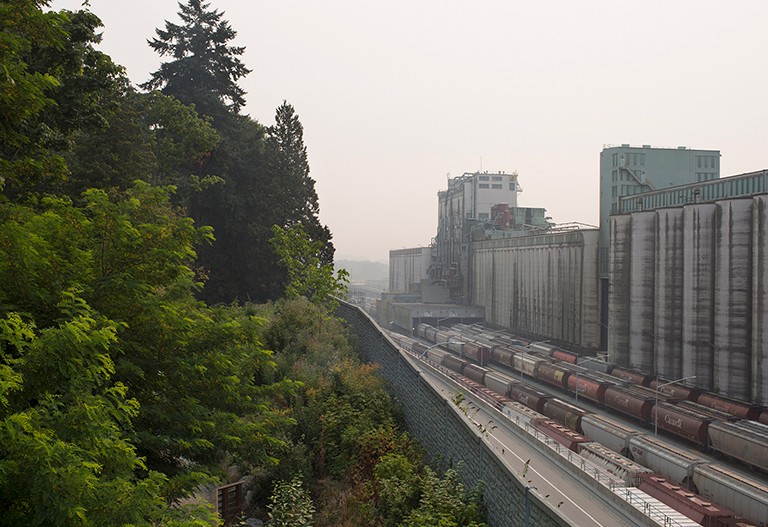 A cityscape shrouded in wildfire smoke, with trees on one side and an industrial area on the other.