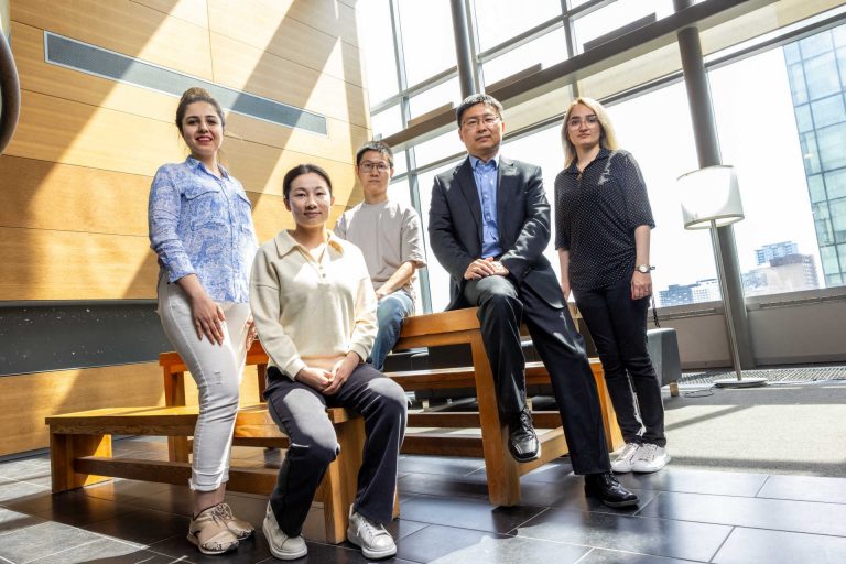 Group photo of 5 people, some sitting and some standing on a wooden table and wooden bench indoors