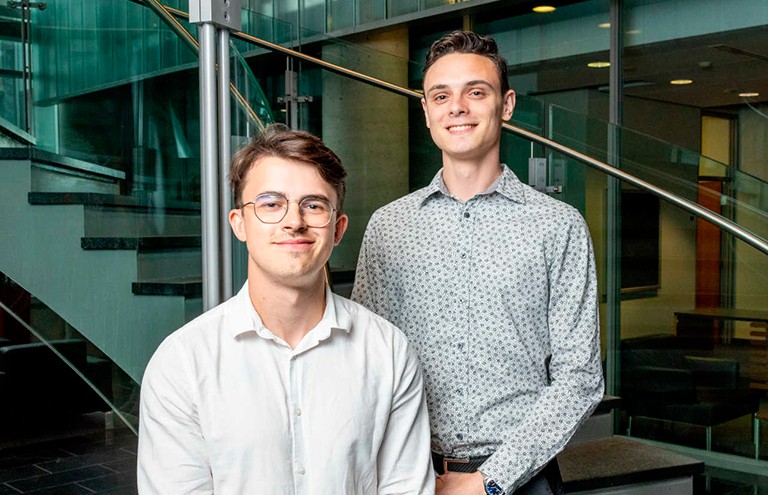 Two smiling young men wearing button down shirts and standing on a stairwell.