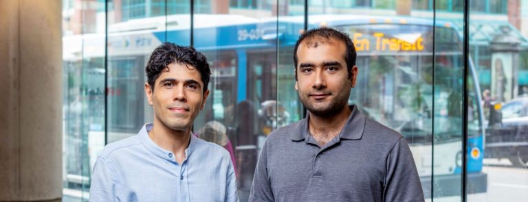 Two men wearing blue shirts stand in front of a ground-floor window with a city bus behind them