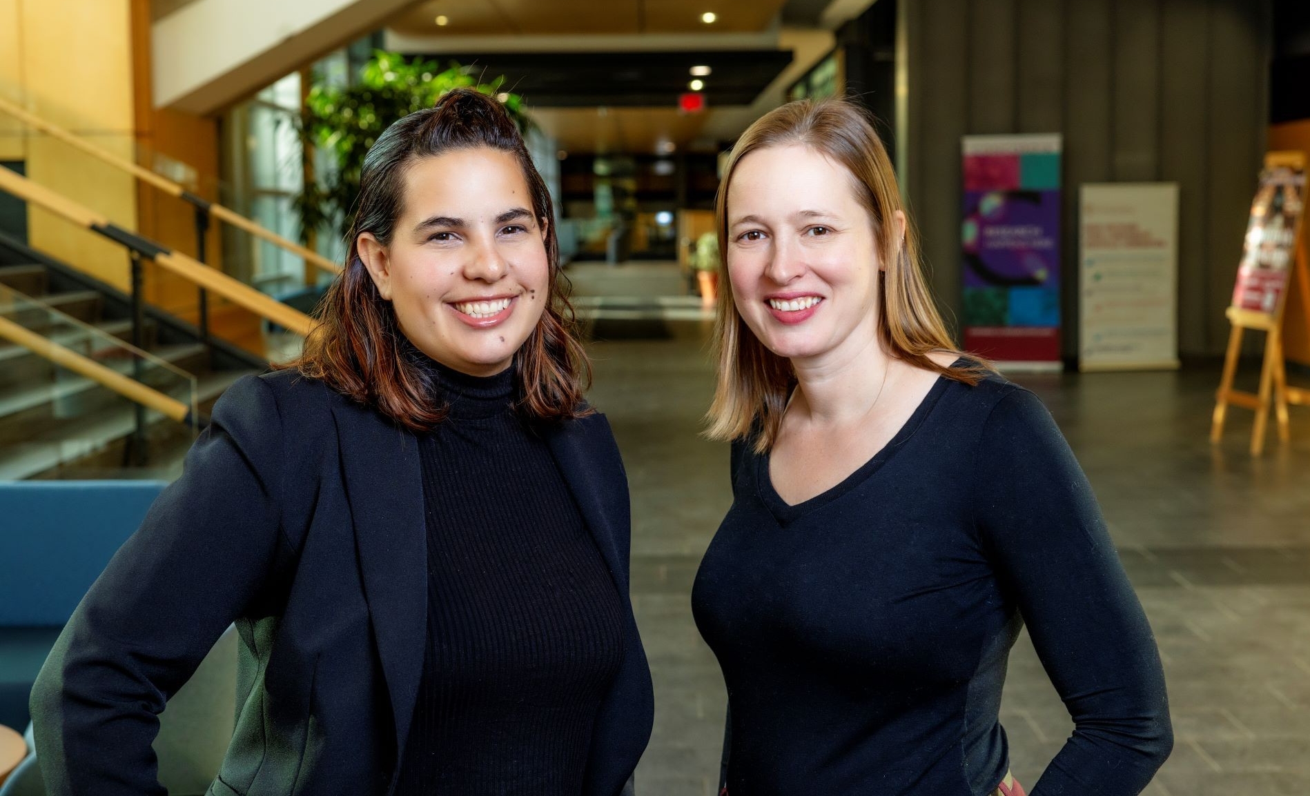Andrea Sander Montant and Krista Byers-Heinlein Two women dressed in dark clothes smile in a university corridor