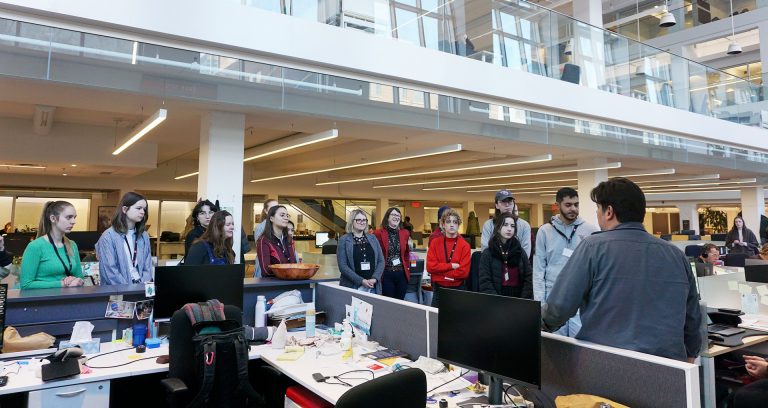 A group of young people being talked to by an older man in a large and light news room