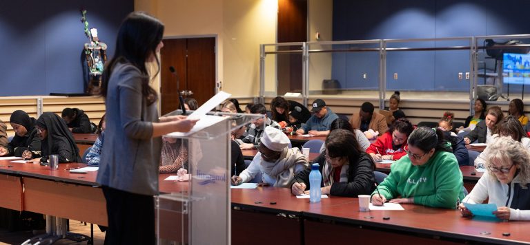 A woman standing at a lectern in a room where people are taking a test.