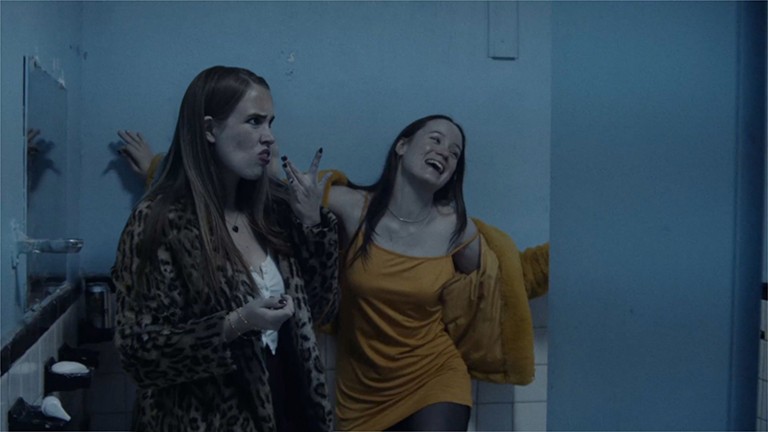 Messy Legends is a love letter to a creative class of artists. Two woman in a public toilet, one scowling, the other laughing uproarously.