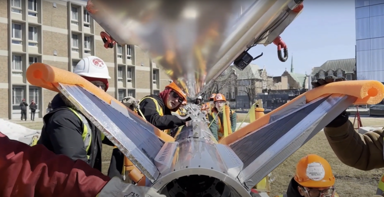 A group of students wearing hard hats and safety gear work on a large metallic rocket-like structure outdoors on a university campus. 