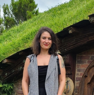 A woman with shoulder-length brown hair, wearing a sleeveless gray vest over a black top, stands smiling in front of a small house with a grass-covered roof and wooden beams. Lush green plants and trees are visible in the background.