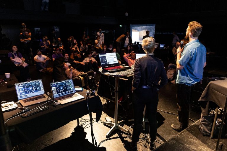 Two presenters stand on a dimly lit stage facing an audience, with multiple laptops and audio equipment set up on tables. The audience, seated in casual arrangements, watches attentively. The presenters, one holding a microphone, appear to be leading a workshop or demonstration involving digital audio or music production software displayed on the laptop screens.