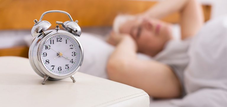 A clock in the foreground with a woman trying to sleep in the background