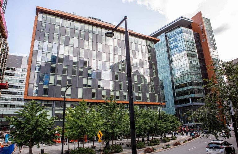 A modern multi-story glass building with reflective windows stands beside a tree-lined street with cars and pedestrians. 