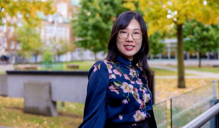 A woman with dark hair in a floral shirt and glasses stands outside in a green area