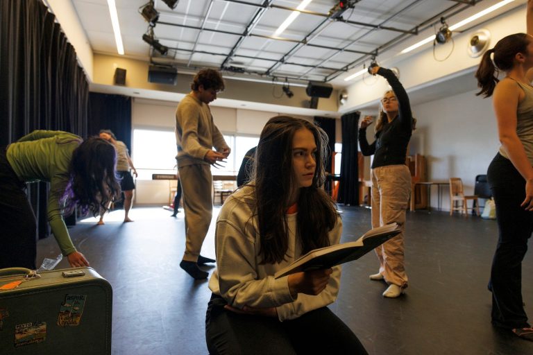 A woman sits on the floor of a studio with a book, surrounded by performers.