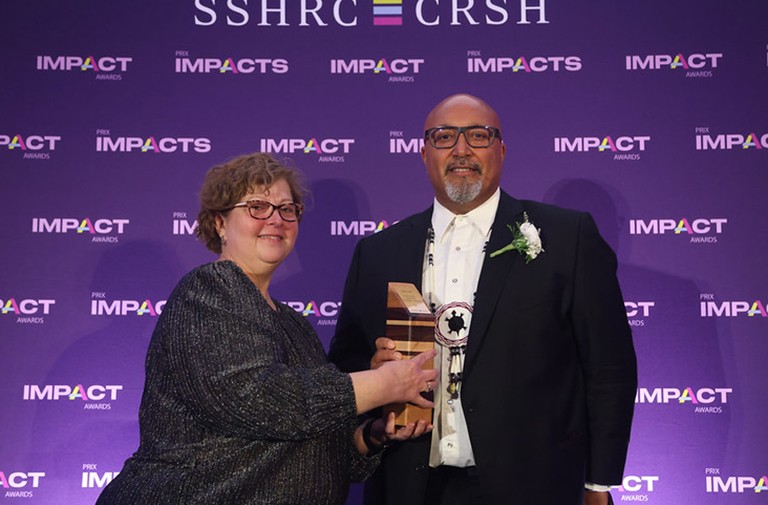 Two smiling people, one handing the other some kind of wooden statue award