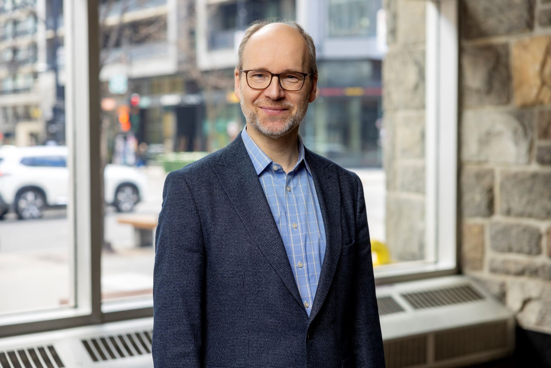 A man  in a blue shirt and blazer stands in front of a large window
