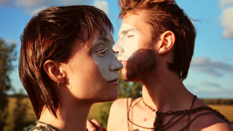 Two young adults in profile stand close together outdoors at golden hour, both wearing white face paint across their eyes and upper faces, with warm sunlight illuminating their skin against a blurred rural