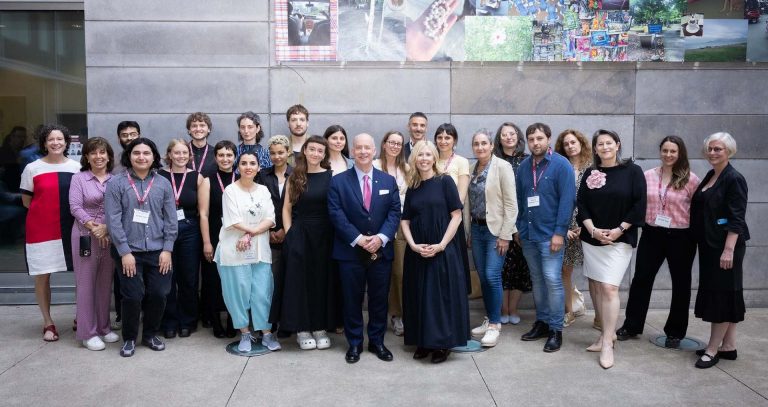 A large group of people standing together and smiling for the camera in a large, concrete interior space with a large artwork hanging in the background
