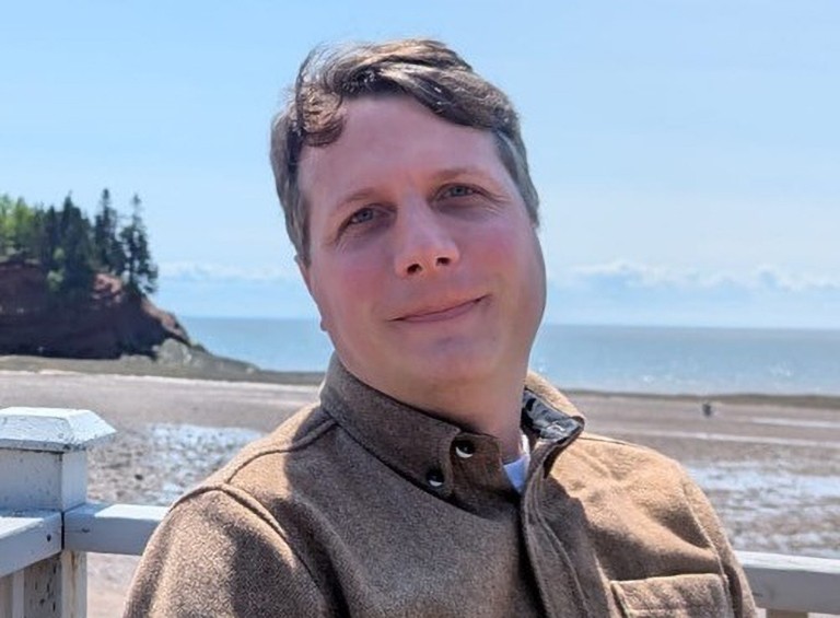 Smiling man standing outdoors with a coastal area at low tide behind him.