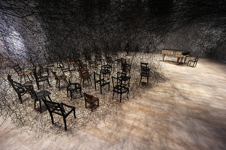 Exhibition view of chairs in a room with dark string woven and coming down from the ceiling