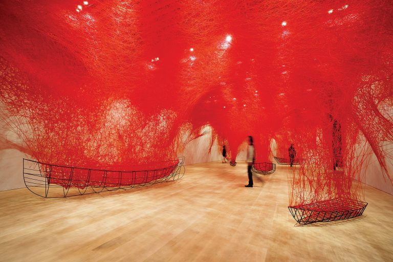An art exhibition with red interwoven strings stretching from the ceiling to the floor, with the skeletons of canoes made from wire resting on the ground