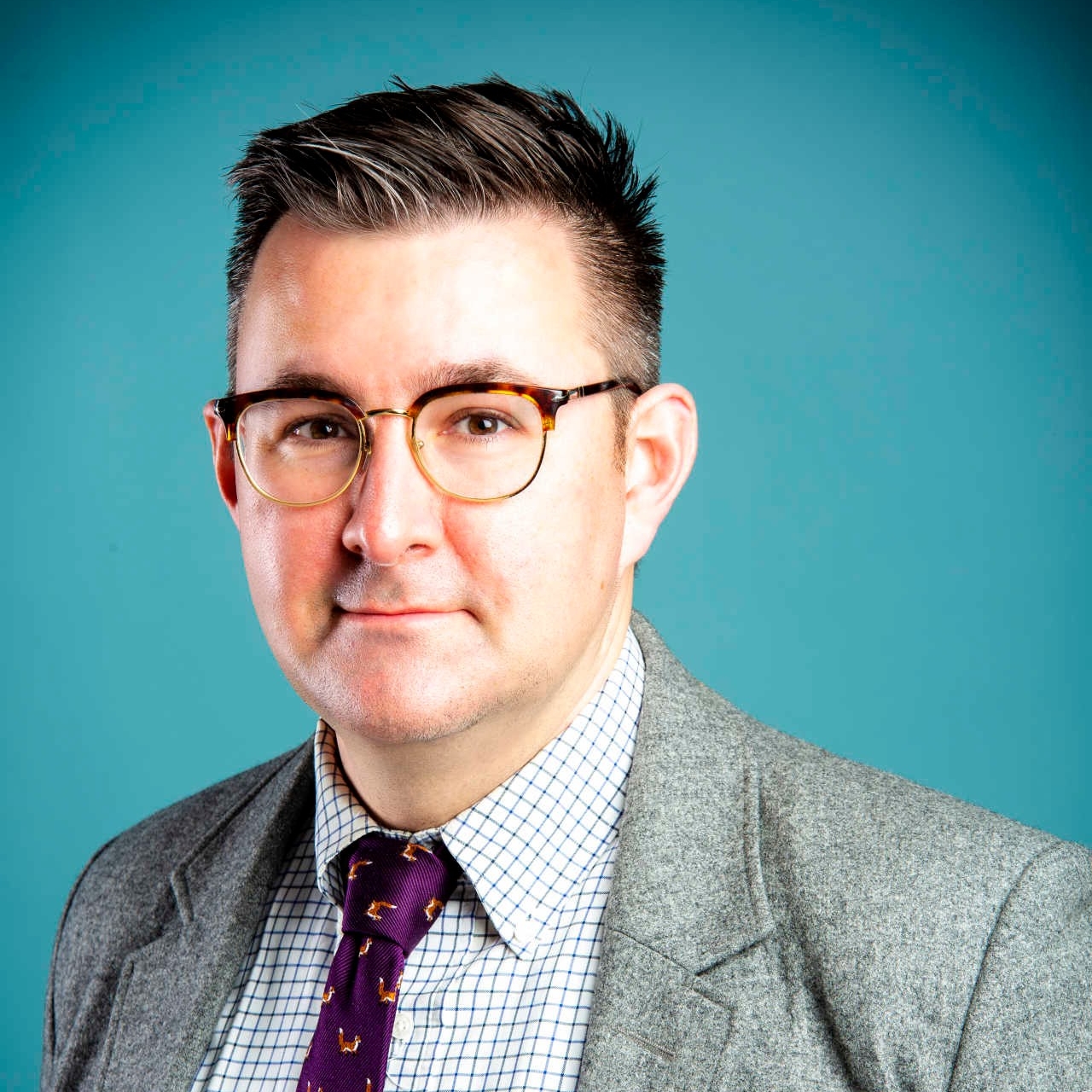 Fenwick McKelvey Fenwick McKelvey pose devant un fond bleu. Il porte une veste grise, une chemise à carreaux bleus et blancs, une cravate violette et des lunettes