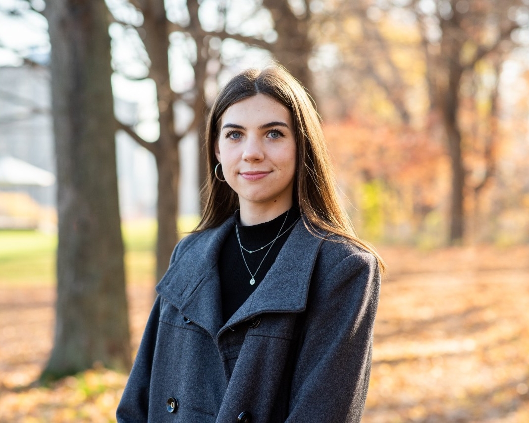 Chloe Cull Chole Cull est prêt à l'extérieur devant des arbres par une journée ensoleillée. Elle porte un manteau bleu et un col roulé noir